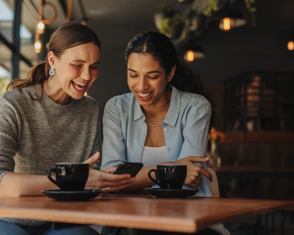 Two friends laughing together over coffee at a sunlit cafe table, phone in hand